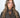 Modell med Small Triangle Scarf Brown fra Sui Ava – lite brunt strikket trekantskjerf knyttet tett rundt halsen.