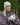 Modell utendørs med MSCH Copenhagen Hope Icon Mittens i lys beige – myke, ribbestrikkede votter med oppbrettede mansjetter og liten logolapp. Modellen holder en lys keramikkopp mellom hendene og har på seg matchende beige strikkelue og en brun, quiltet jakke.