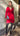 Modell i A-View Carrie Dress Red – kort, rød omslagskjole med glansfull satenglook, V-utringning, lange ermer, knyting i midjen og asymmetrisk skjørt med volanger. Kjolen er stylet med sorte strømpebukser, burgunder pumps og en sølvfarget paljettveske.