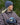 Modell utendørs med MSCH Copenhagen Hope Icon Beanie i melert blå, en myk strikkelue med bred oppbrettet kant og hvit logolapp foran. Luen er stylet med matchende blå strikkevotter og en brun, quiltet jakke. Modellen holder en blå keramikkopp mellom hendene.